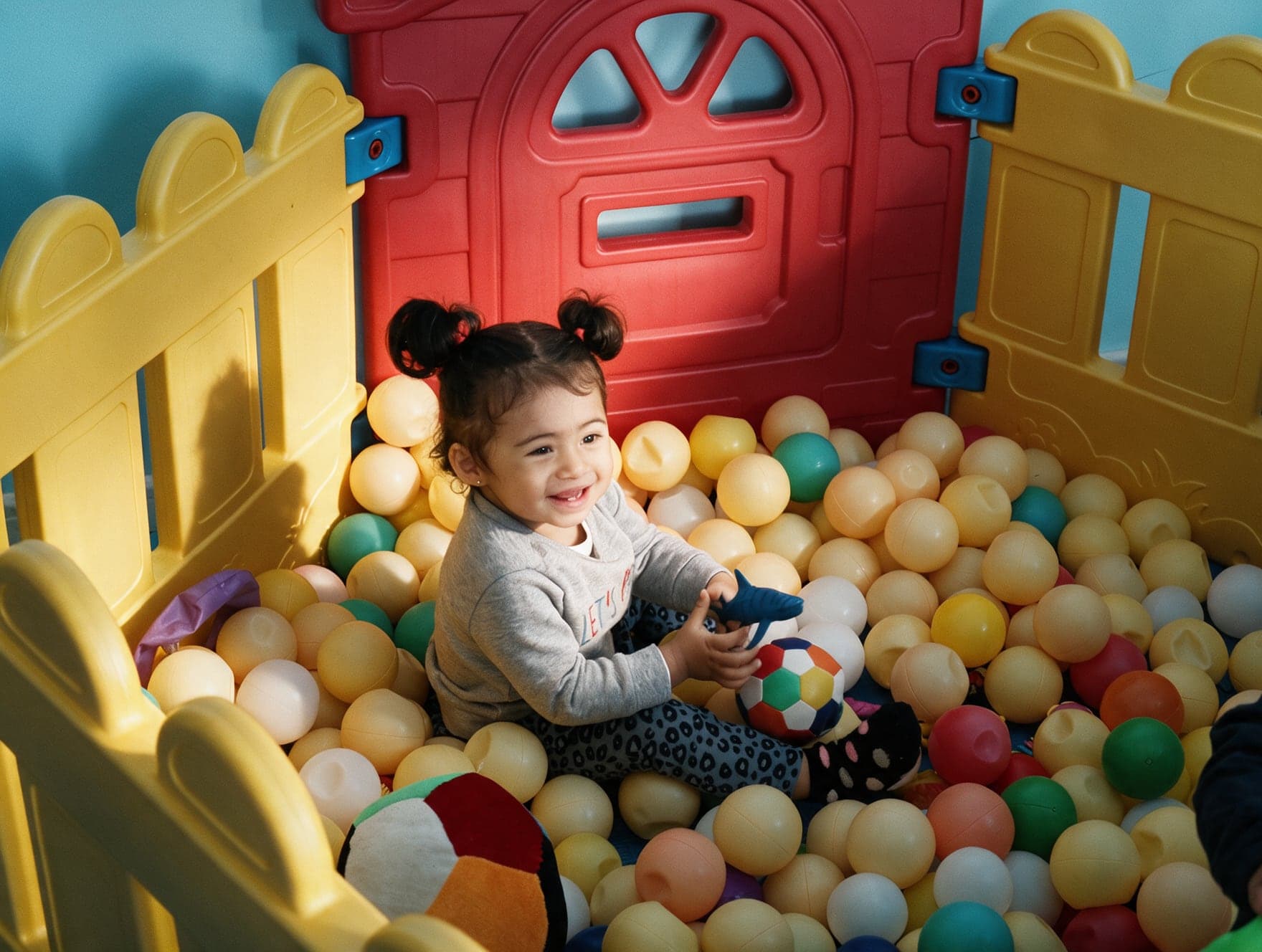 A toddler laughing in the ball pit inside a yellow and red play pen